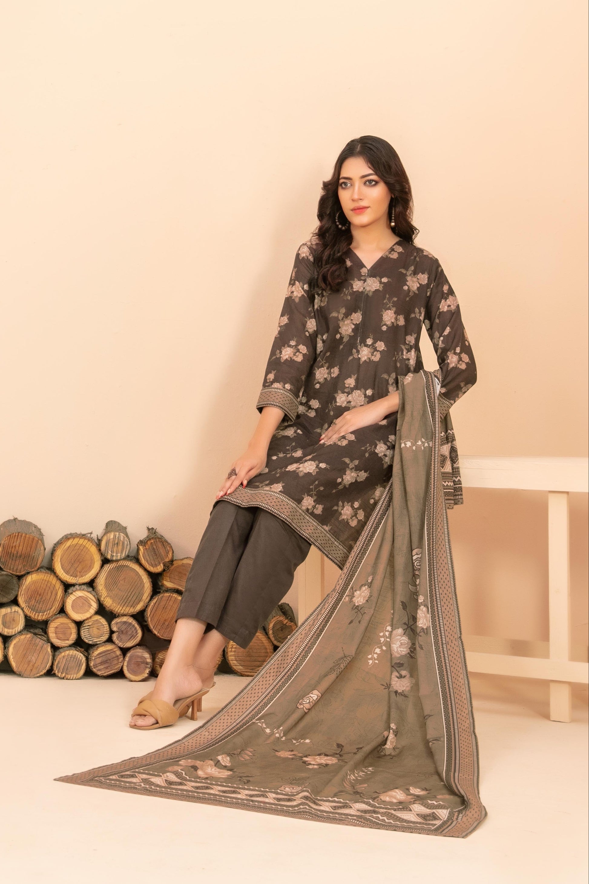 Woman in a floral dress standing next to stacked wooden logs against a beige wall.