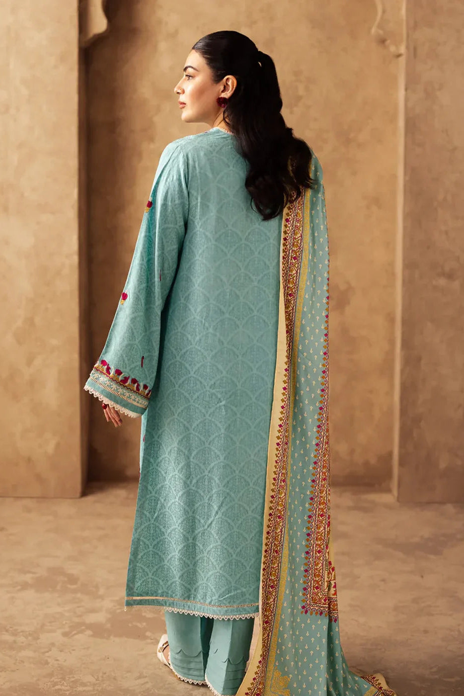 Woman wearing a light blue traditional outfit with a long, embroidered scarf against a beige wall.