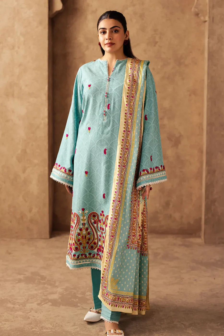 Woman wearing a traditional outfit with a light blue top and patterned dupatta against a beige wall.