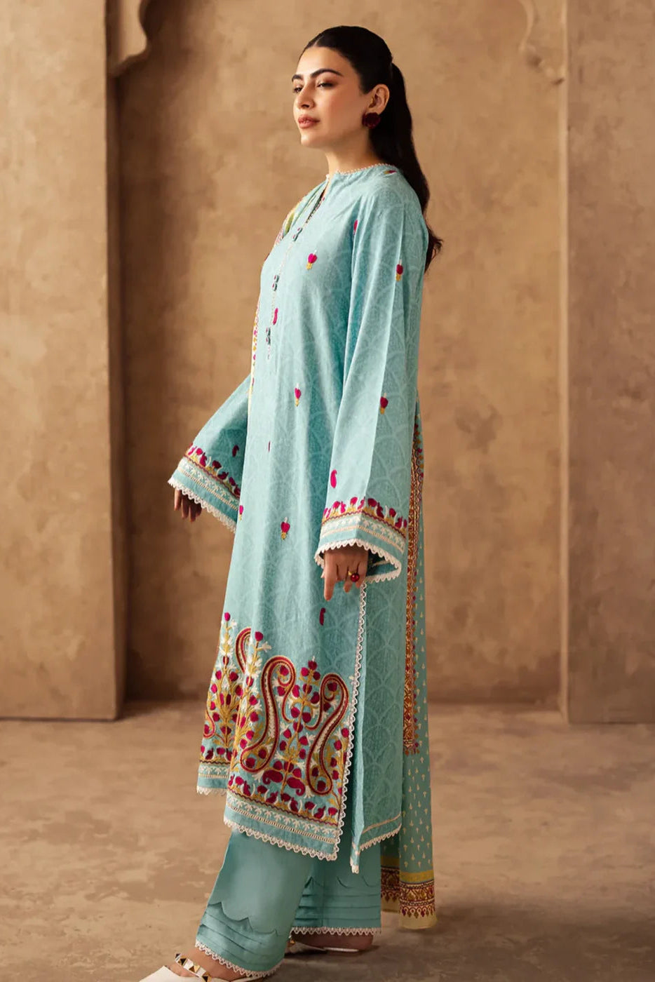 Woman wearing a light blue embroidered traditional outfit against a beige wall.