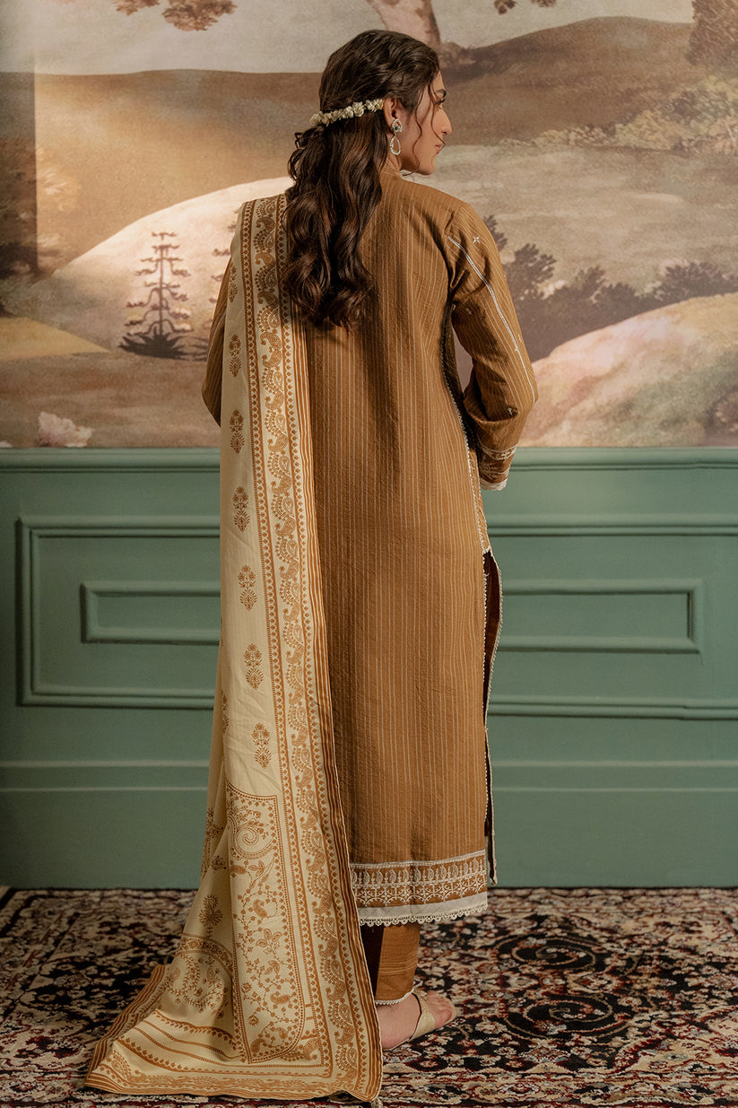 Woman in a brown traditional outfit with a patterned scarf, standing in front of a decorative wall.