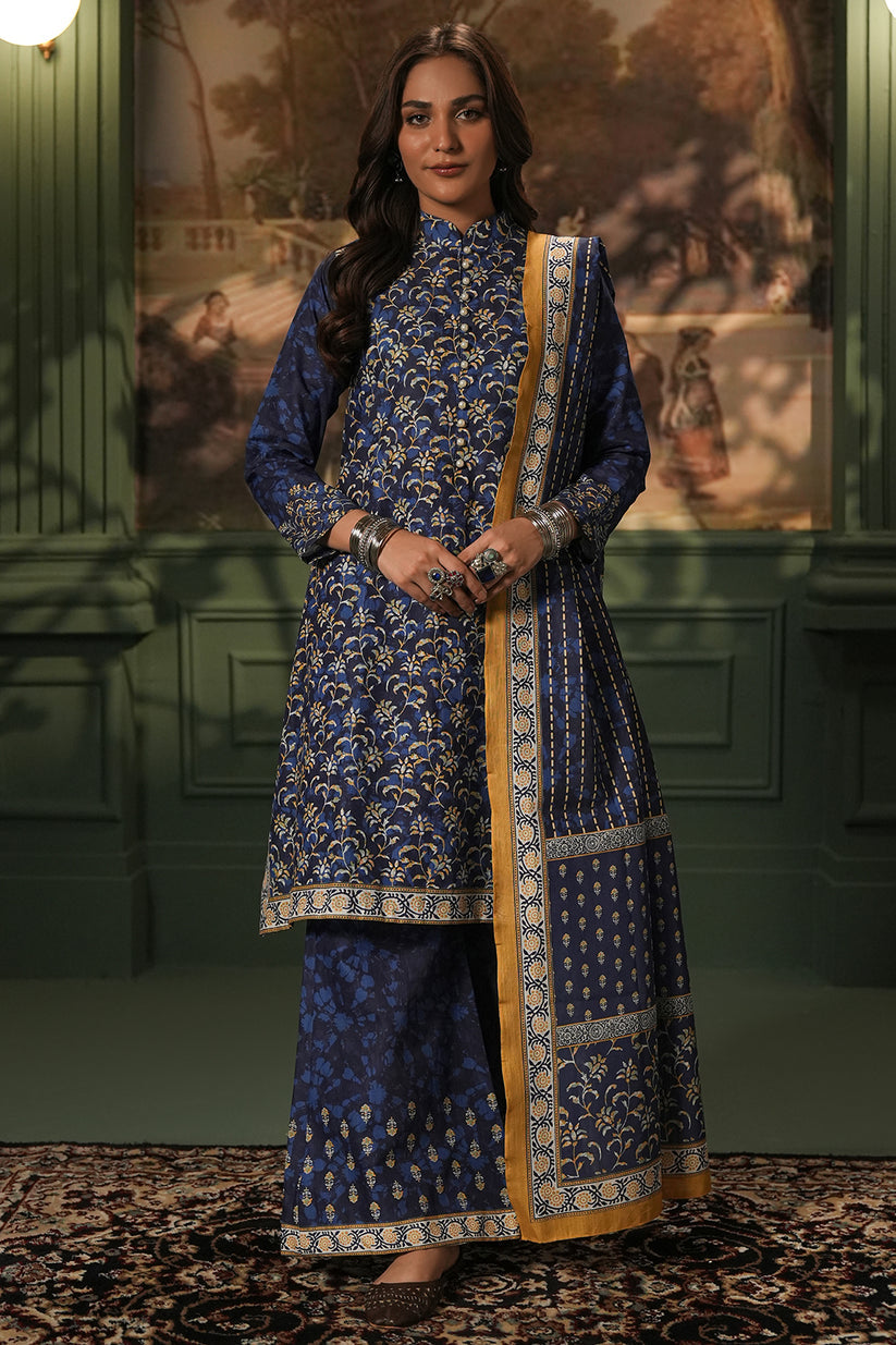 Woman wearing a traditional blue and gold outfit with a decorative border against a green wall.